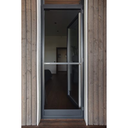 Tinklelis nuo uodų - varstomos durys su metaliniu rėmu yra iš dalies atidarytas medinėmis dailylentėmis apkaltoje sienoje, matosi medinės grindys.