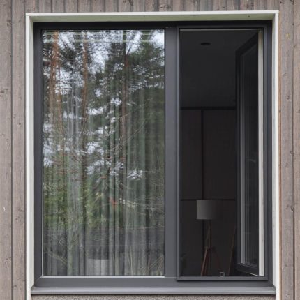 Didelis langas su Tinklelis nuo uodų - langams su rėmeliais ir vertikaliomis žaliuzėmis, vienas stiklas atidarytas, jame atsispindi medžiai ir lempa.