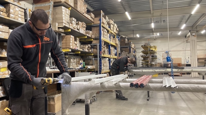 Divi strādnieki montē iekārtas insektu sietu ražošanai noliktavā, kuru ieskauj plaukti ar kastēm un materiāliem.